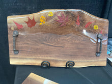 Load image into Gallery viewer, Live Edge Walnut, rich browns and warm golds, clear food safe epoxy covering 30% of wood with real dried leaves and flowers in the epoxy, textured aged iron handles.
